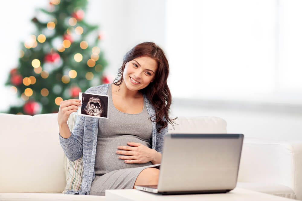 Happy Pregnant Woman With Laptop Computer Having Video Call and Showing Ultrasound Image Over Christmas Tree Background