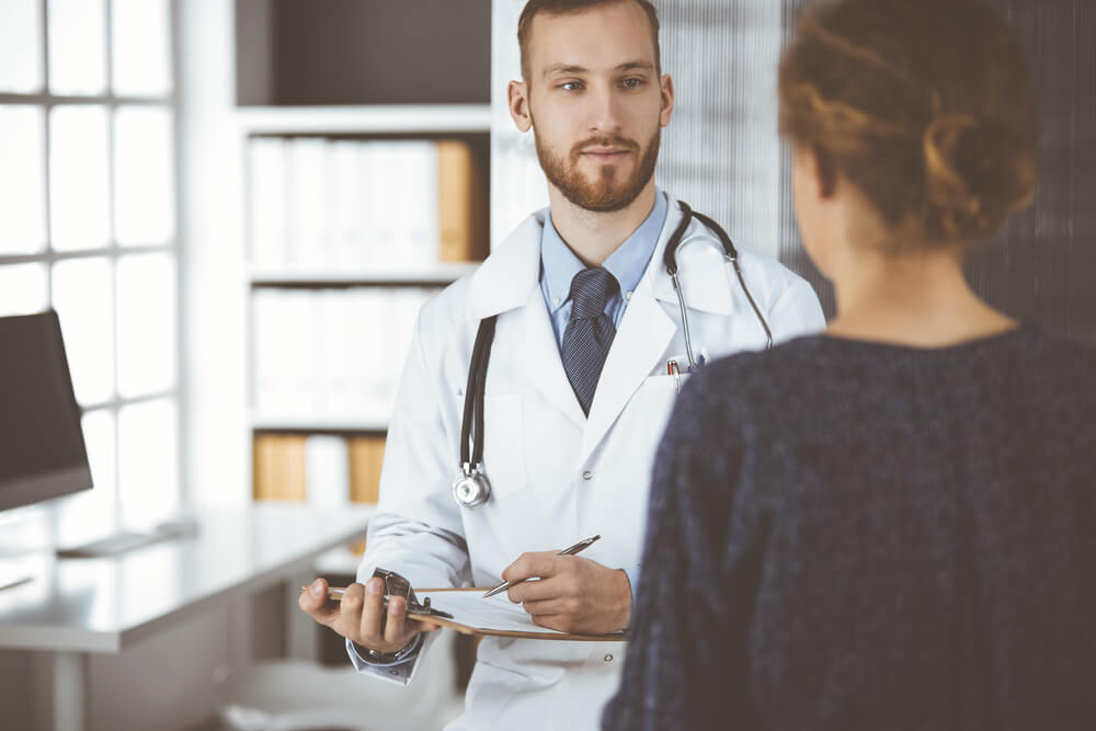 Friendly Red-Bearded Doctor and Patient Woman Discussing Current Health Examination While Sitting in Sunny Clinic.