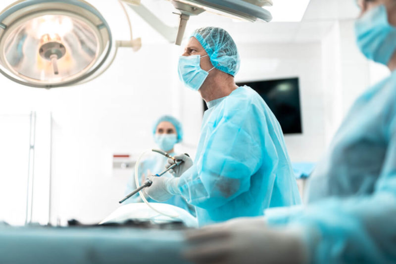 Side View Portrait of Focused Doctor in Sterile Gloves Holding Laparoscopic Instrument While Looking Away With Serious Expression.