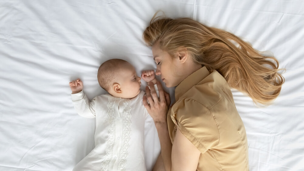 Tired Mom and Cute Peaceful Baby Sleeping on Mattress With White Sheet. 