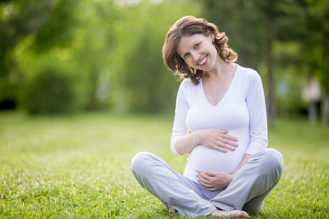 Portrait of Happy Young Pregnant Model Sitting With Crossed Legs on Grass Lawn and Looking at Camera.
