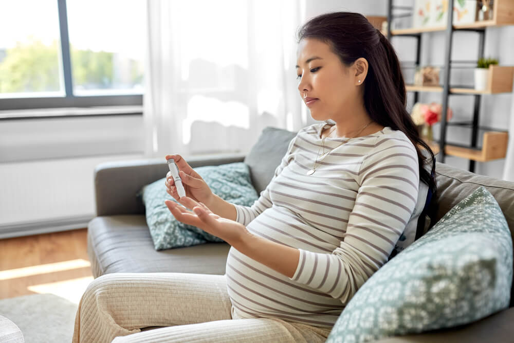 Pregnant Asian Woman Checking Blood Sugar Level With Glucometer and Lancing Device at Home