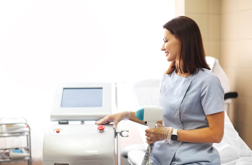 Young Woman Beautician in Medical Uniform Adjusting Machine for Photorejuvenation or Laser Hair Removal