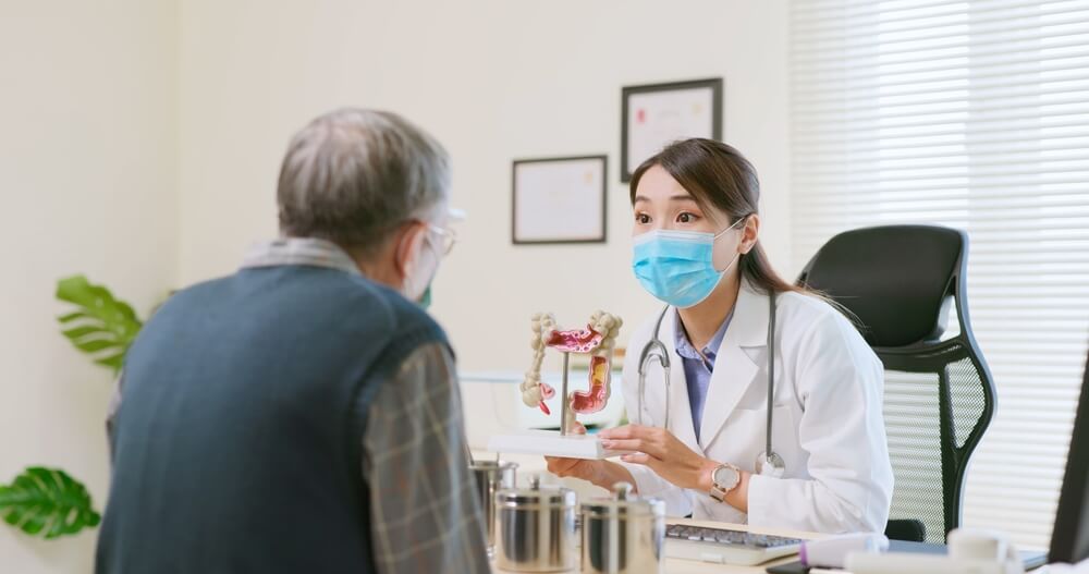 Asian Female Doctor Wearing Face Mask Is Showing a Model of the Large Intestine and Explaining to Elder Senior Man Patient in Hospital