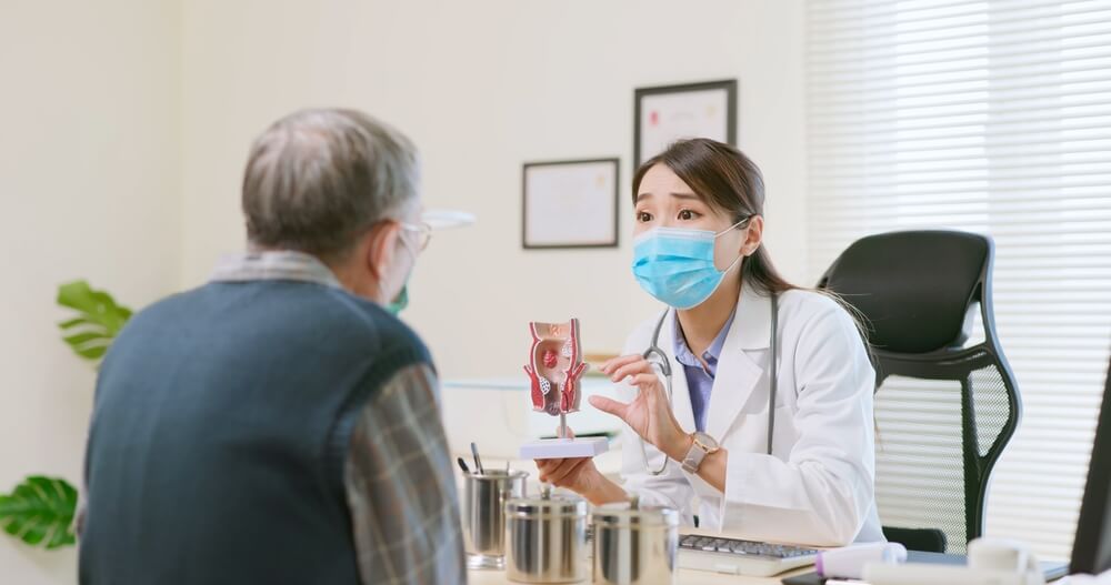Asian Female Doctor Wearing Face Mask Is Showing A Rectal Model And Explaining To Elder Senior Man Patient In Hospital