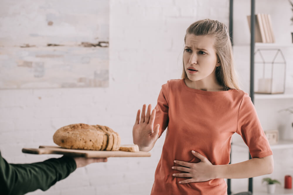 Man Holding Cutting Board With Bread Near Upset Blonde Woman With Gluten Allergy