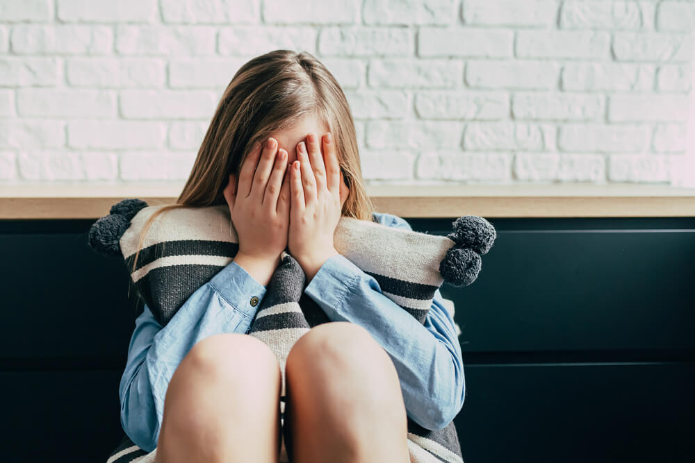 Sad Caucasian Girl in Depression Hugs a Pillow and Closes Her Eyes With Her Hands on a Background of a White Brick Wall.