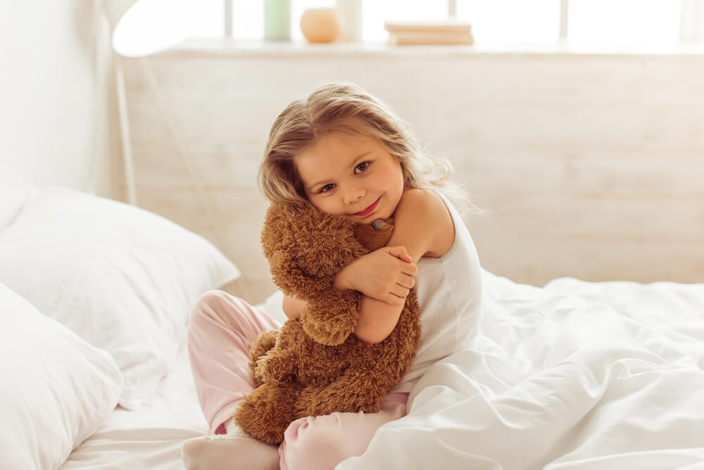 Sweet Little Girl Is Hugging a Teddy Bear, Looking at Camera and Smiling While Sitting on Her Bed at Home