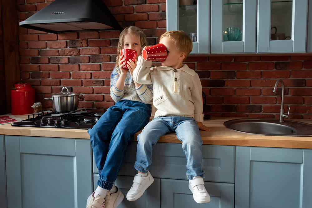 Pair of Cute Adorable Caucasian Blond Siblings Boy and Girl Enjoy Have Fun Sitting on Kitchen Counter and Drinking Hot Tea