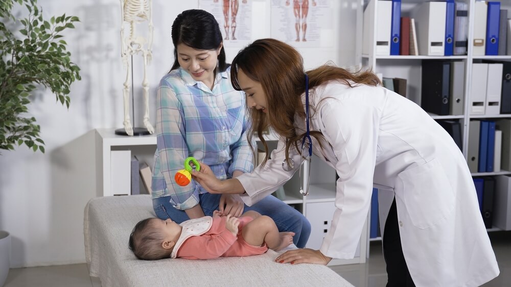 Asian Pediatrician Stooping Over the Baby on Treatment Couch.