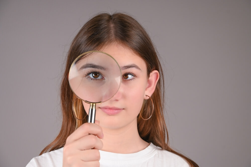 Teenage Girl With Magnifying Glass on Gray Background. Female Eyes With Strabismus.