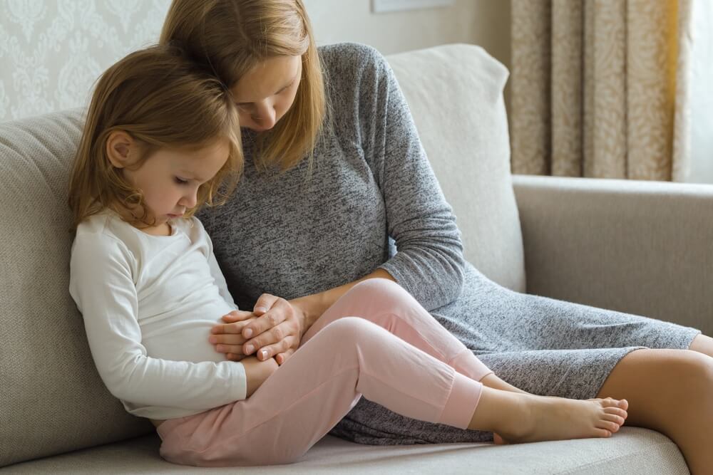 Child Sitting On Sofa And Suffering From Stomach Ache At Home Room.