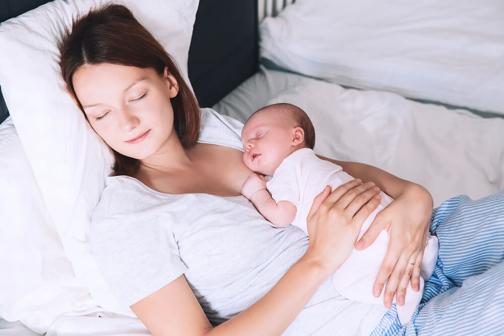 Newborn Baby Sleeping In The Hands Of His Mother. Image Of Happy Maternity And Co-sleeping. Mom And Child's First Month Of Life At Home.