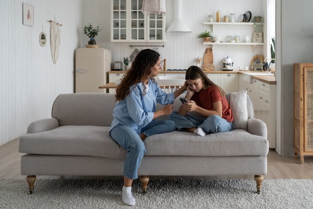 Disappointed teen girl looking at phone screen, sharing feelings with her mother.