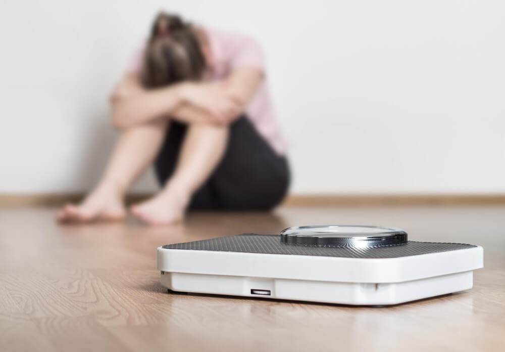 Depressed, Frustrated and Sad Woman Sitting on Floor Holding Head and Arms on Knees Behind a Weight Scale
