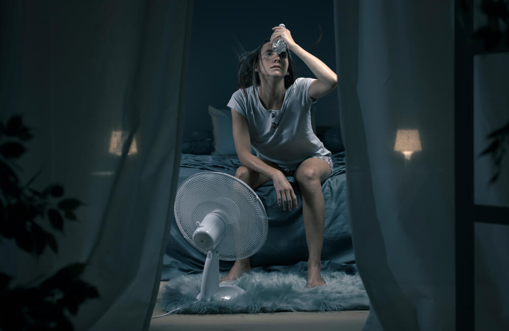 Exhausted Woman Suffering Suring the Heatwave, She Is Holding a Water Bottle and Sitting in Front of a Cooling Fan in the Bedroom