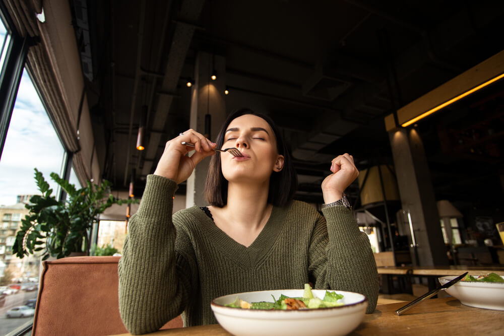 Satisfied Young Woman Enjoys Tasty Salad in a Restaurant, Putting Fork Into Her Mouth and Closing Her Eyes.