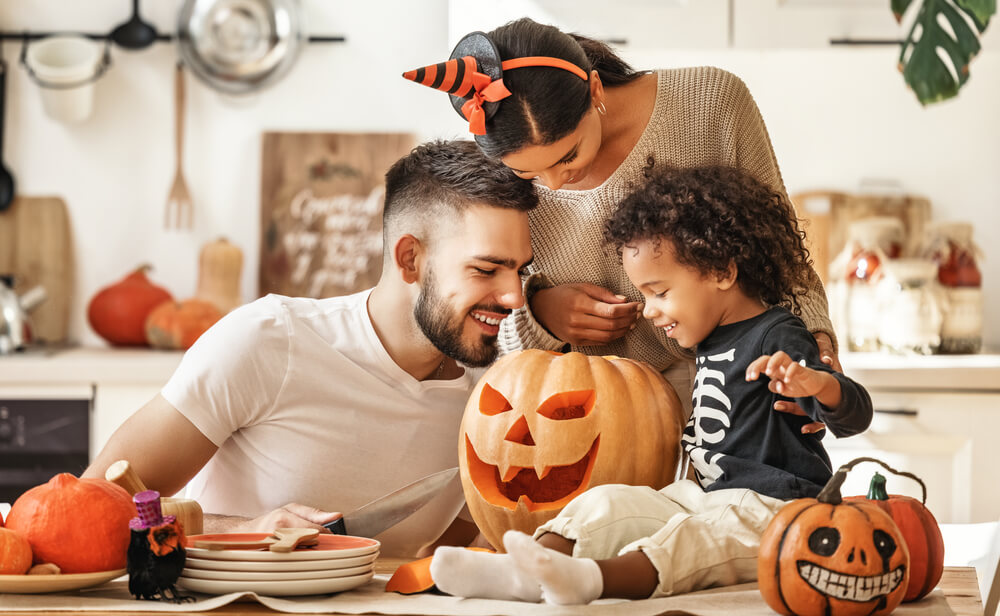Cheerful family - parents with son smiling while creating jack o lantern from pumpkin during Halloween celebration in kitchen at home