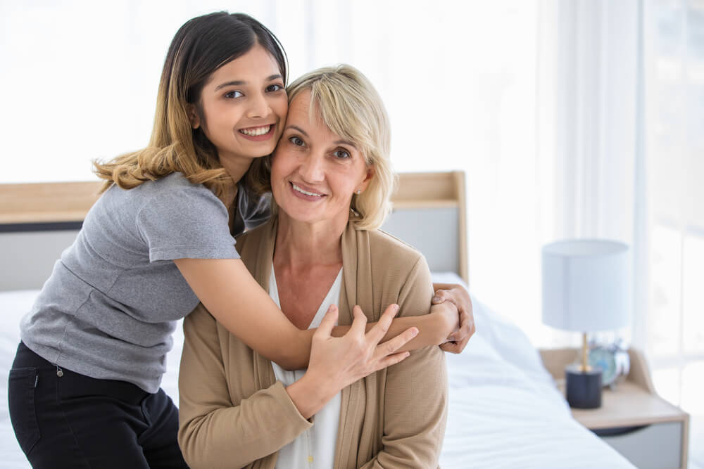Middle Aged Woman Hugging Gently Cheerful Asian Adopted Daughter and Smiling Happily While Enjoying Time Together at Home