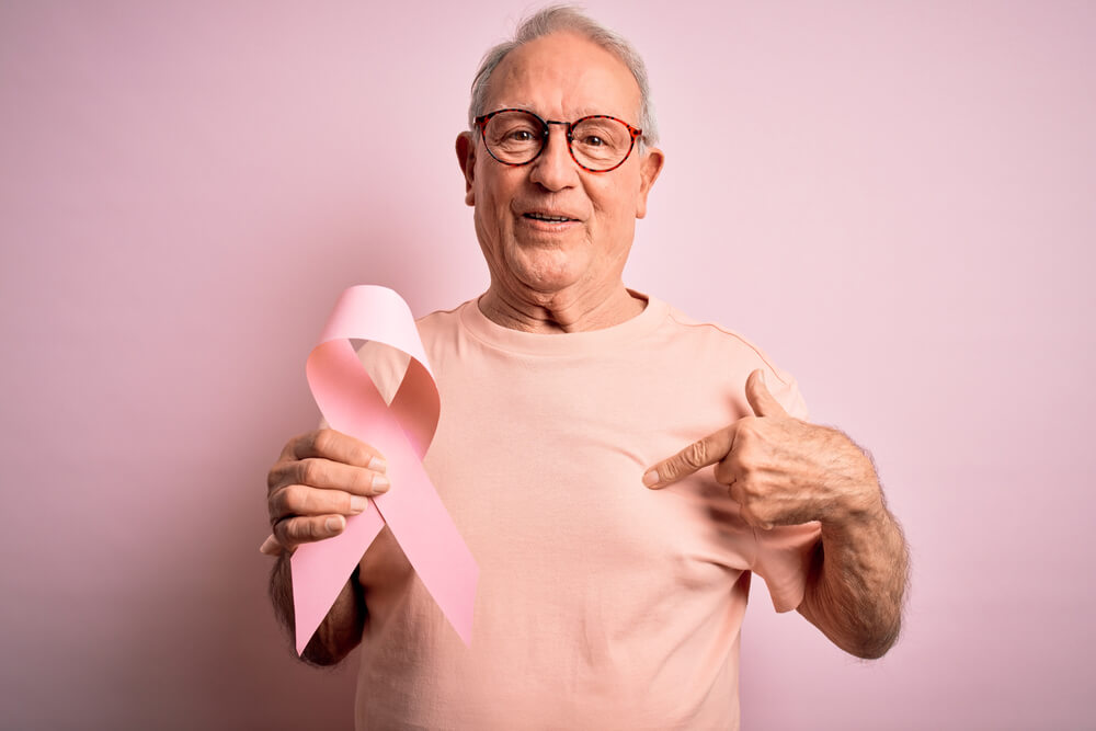 Grey Haired Senior Man Holding Breast Cancer Awareness Pink Ribbon Over Pink Background With Surprise Face Pointing Finger to Himself