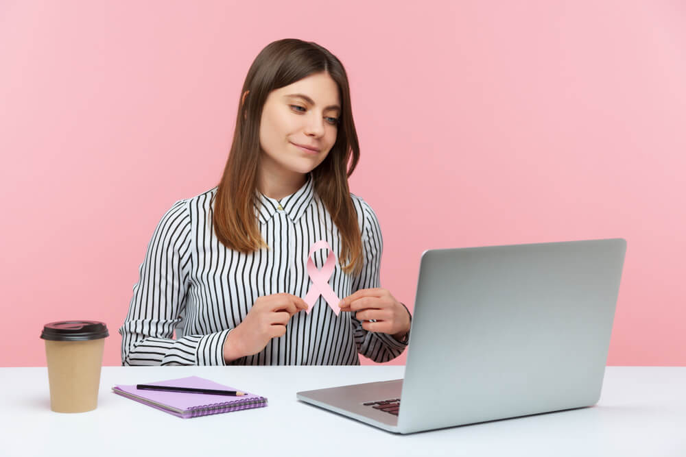 Optimistic Woman Office Worker Holding Pink Ribbon Talking on Video Call at Laptop