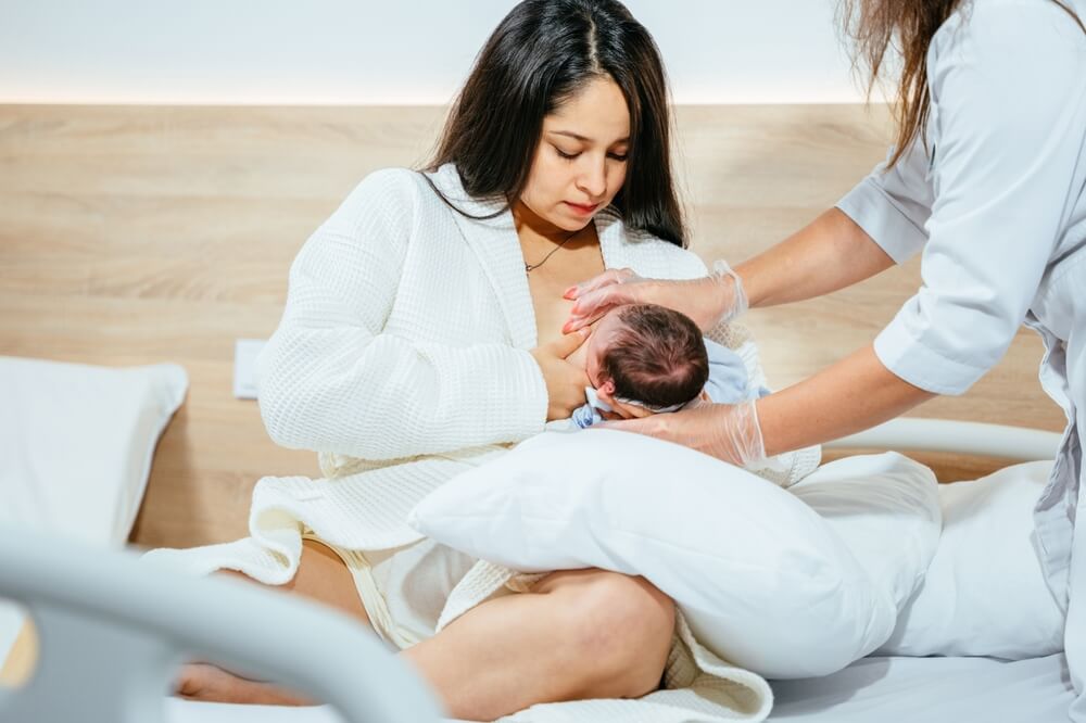 A young mother breastfeeding newborn baby in hospital ward, breastfeeding specialist or nurse help her.
