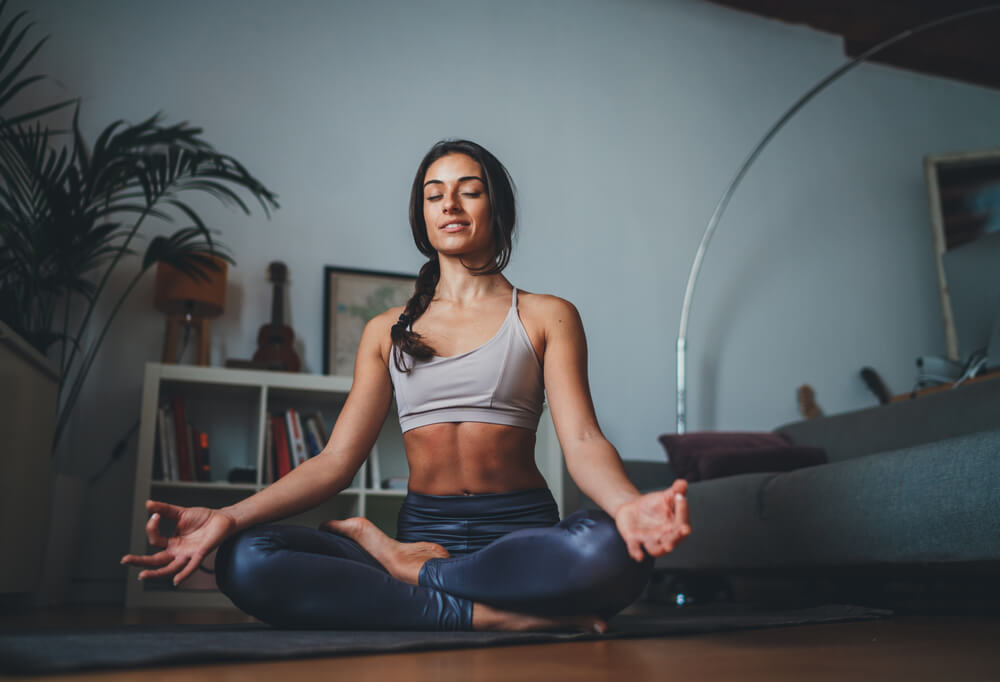 Young Healthy Beautiful Woman in Sportive Top and Leggings Practicing Yoga at Home Sitting in Lotus Pose on Yoga Mat Meditating Smiling