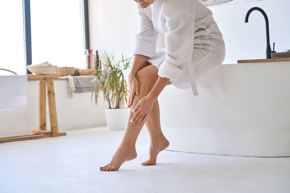 Mid Age Adult 50s Age Mature Woman Applying Varicose Prevention Treatment Cream Massaging Legs Sitting on Bathtub Wearing White Bathrobe.
