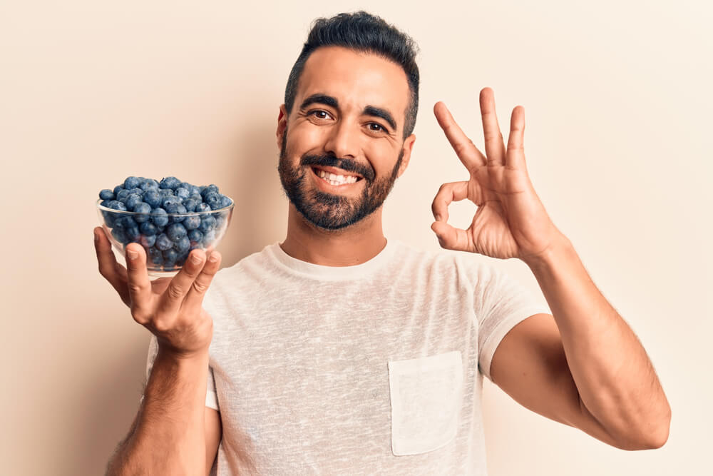 Young Hispanic Man Holding Blueberries Doing Ok Sign With Fingers