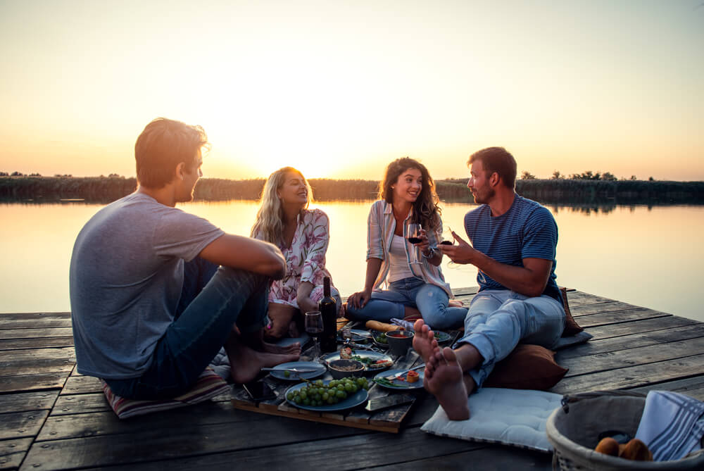 Group of Friends Having Fun on Picnic Near a Lake, Sitting on Pier Eating and Drinking Wine