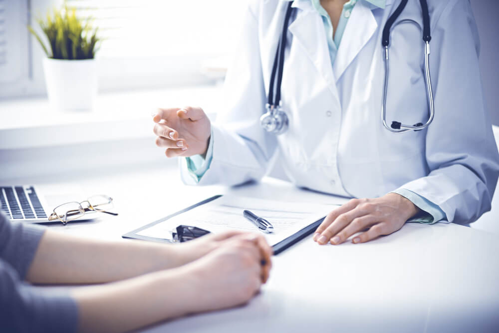 Doctor and Female Patient Sitting at the Desk and Talking in Clinic Near Window.