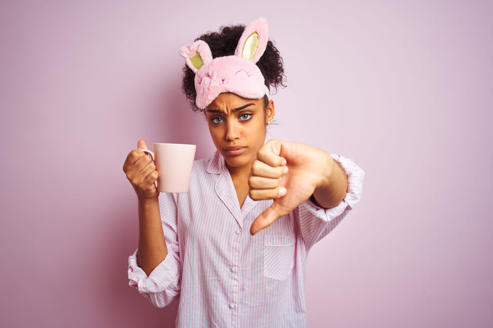 Afro Woman Wearing Pajama and Mask Drinking a Cup of Coffee Over Isolated Pink Background With Angry Face