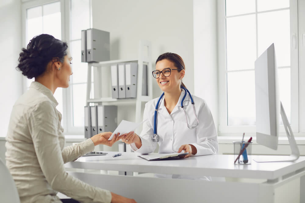 Smiling Female Patient at Consultation With Woman Doctor Sitting at Table in Office Clinic