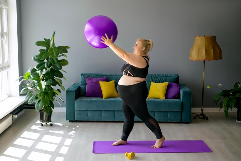 A pregnant plus-size woman is doing fitness at home lifting the fitball up
