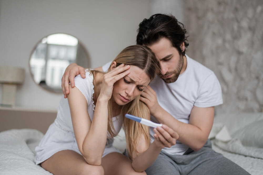 Upset Young Husband Comforting His Depressed Young Wife With Negative Pregnancy Test on Bed at Home.