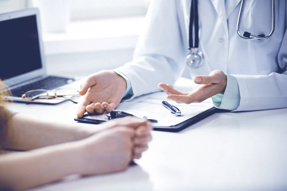 Doctor and Female Patient Sitting at the Desk and Talking in Clinic Near Window