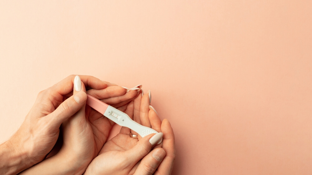 Hands Hold Positive Pregnant Test With Silk Ribbon on Pink Background.