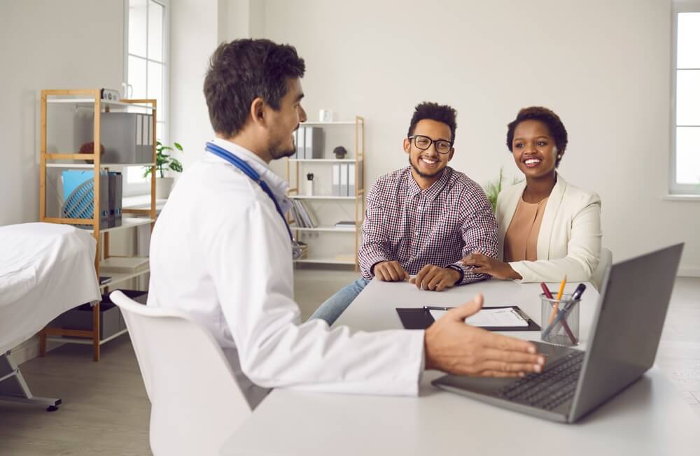 Smiling Male Doctor Consult Excited Multiethnic Family Clients Show Results on Computer in Clinic