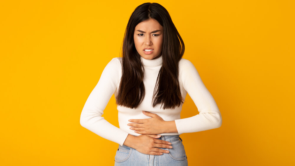 Asian Girl With Stomachache Having Food Poisoning or Menstrual Period Cramp, Yellow Background