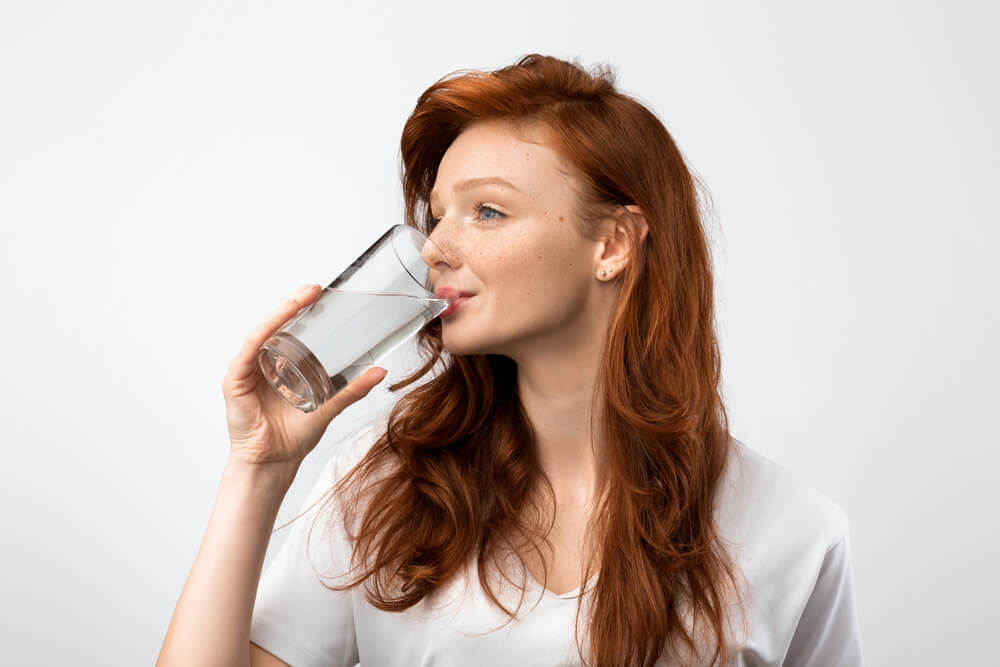 Healthy Hydration. Red-Haired Millennial Woman Drinking Water From Glass Standing Over Gray Studio Background, Wearing Casual T-Shirt