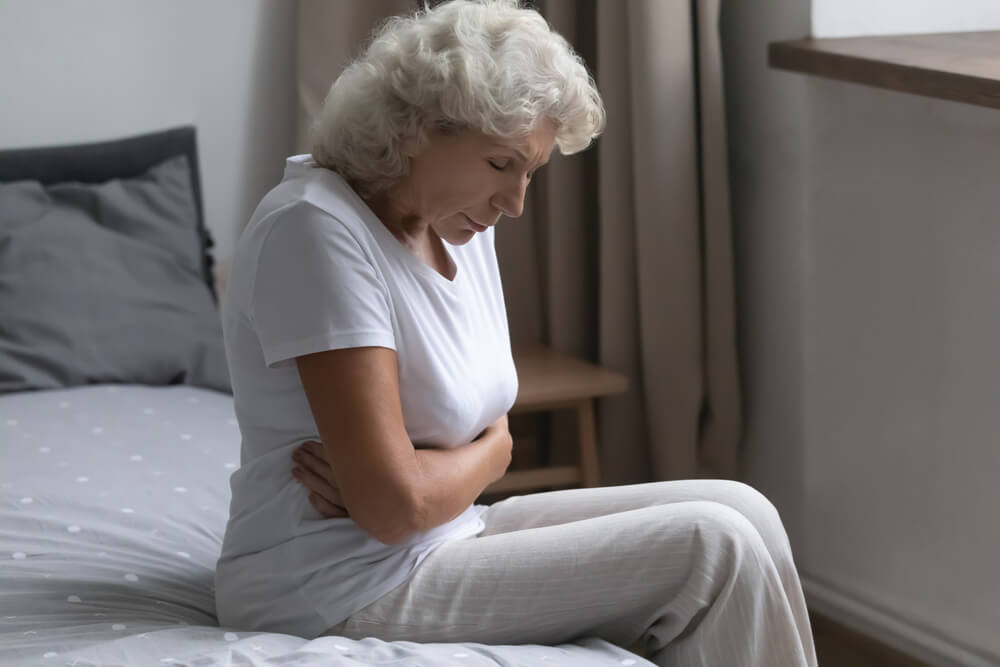 Frustrated Mature Woman Holding Belly, Suffering From Bellyache, Sitting on Bed at Home