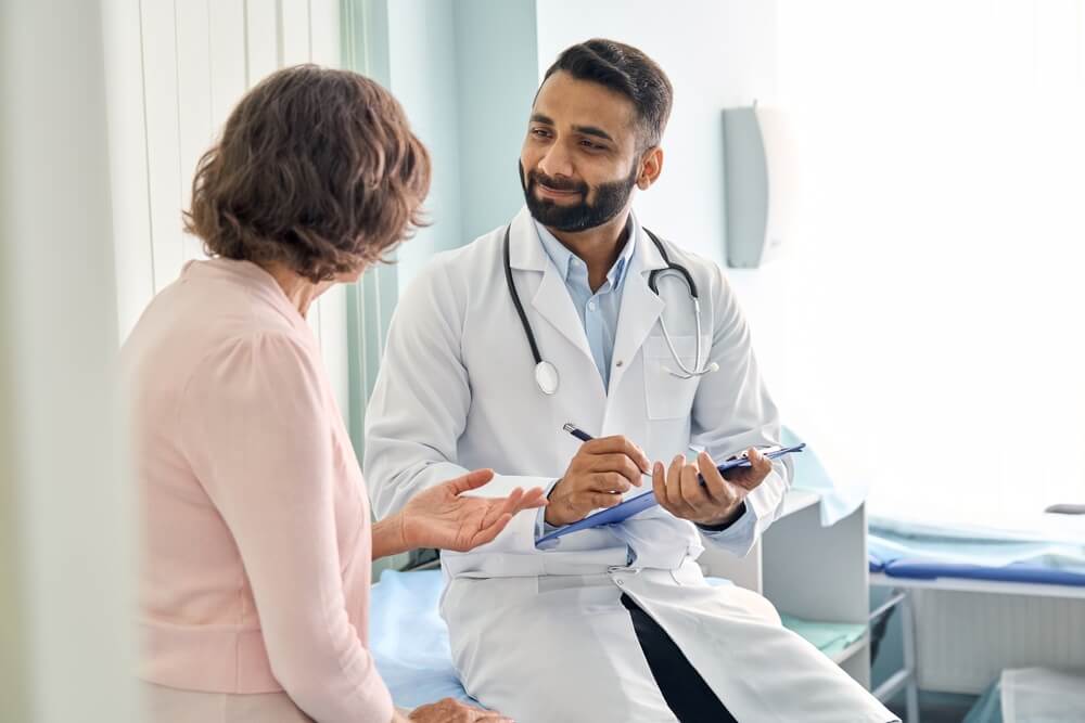 Indian Male Doctor Consulting Senior Old Patient Filling Form at Consultation.