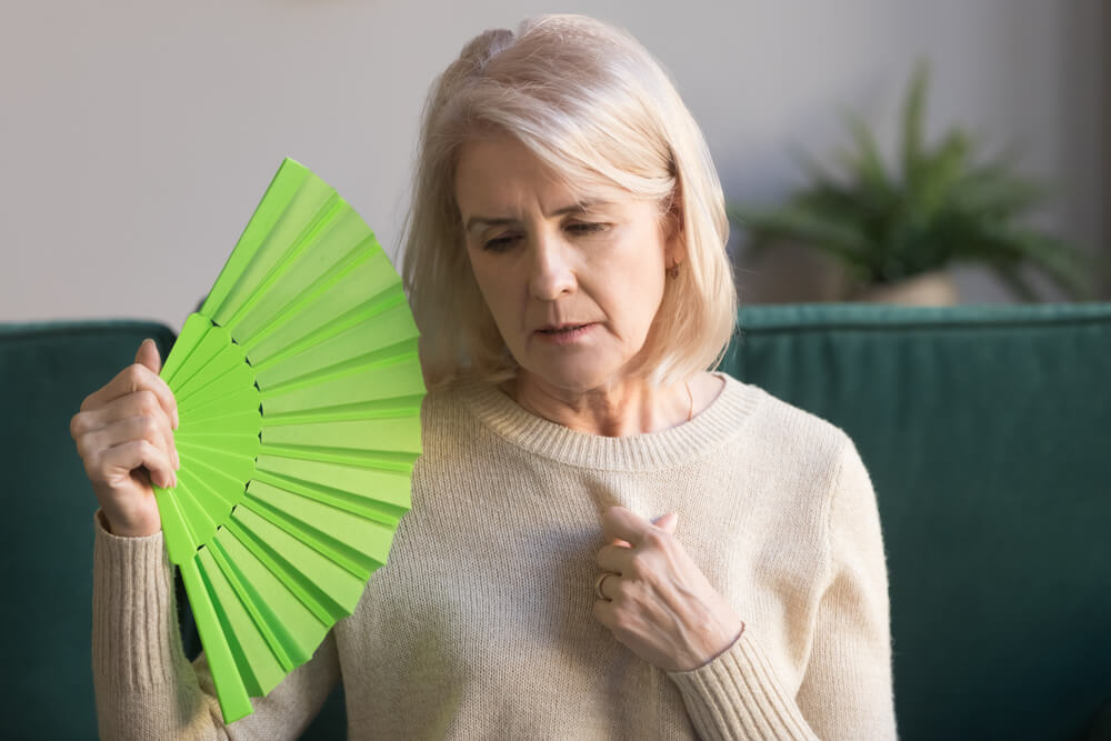 Overheated Elderly Woman Sitting on Couch Waving Green Fan to Cool Herself