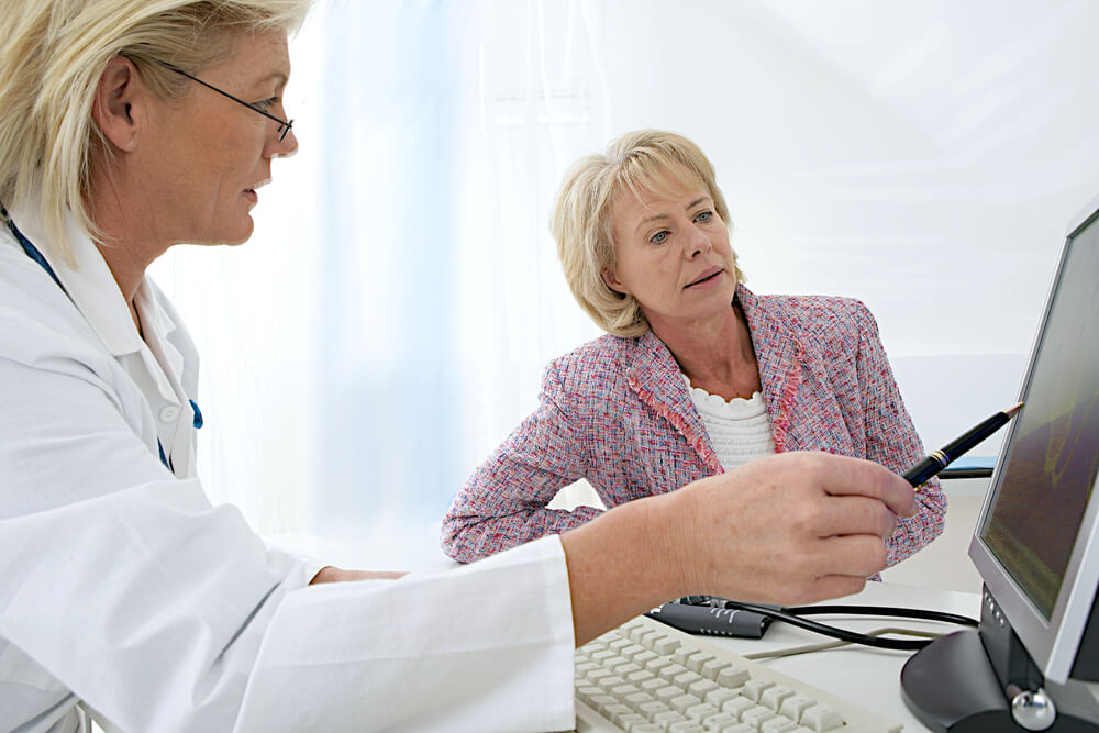 Female Doctor Sitting at Her Desk Pointing to a Computer Screen Explaining Something to a Senior Woman Patient