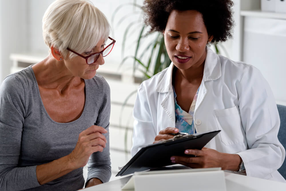 A Female Doctor Sits at Her Desk and Chats to an Elderly Female Patient While Looking at Her Test Results