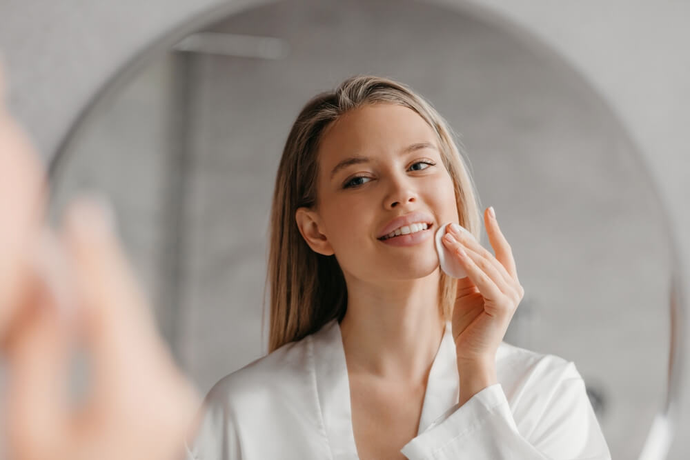 Young Woman Caring for Facial Skin Using Cotton Pad and Looking in Round Mirror in Bathroom Interior.