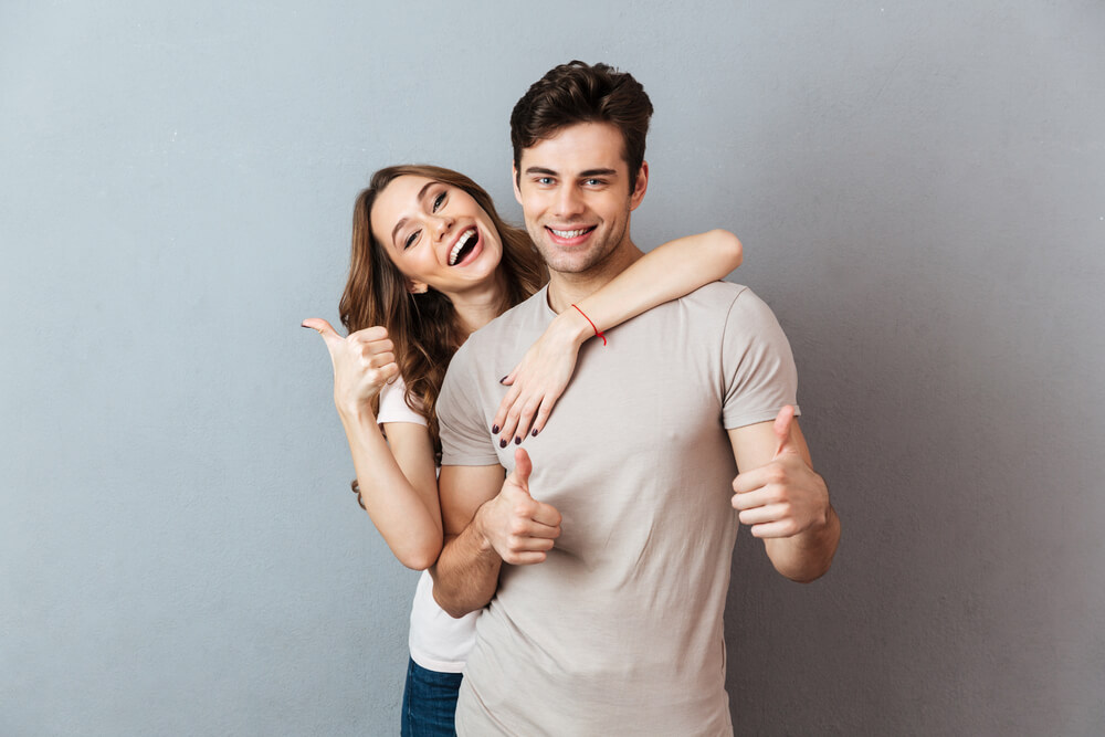 Portrait of a Happy Young Couple Hugging While Standing and Showing Thumbs up Gesture Over Gray Wall