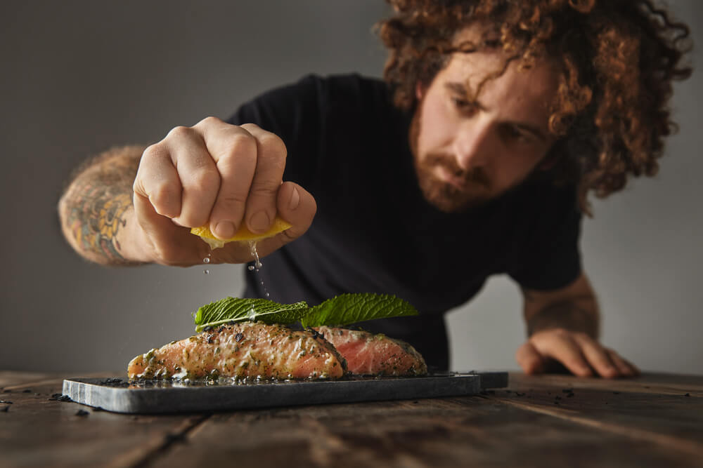 Man Cooks Healthy Meal, Squeezes Lemon on Two Raw Pieces of Salmon Decorated With Mint Leaf in White Wine Sauce With Spices and Herbs Presented on Marble Deck Prepared for Grill