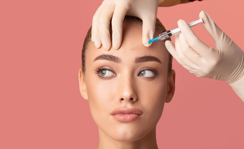 Young Woman Receiving Botox Beauty Injection in Forehead Standing Over Pink Background