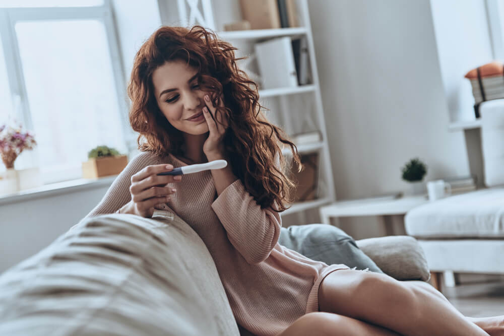 A young women looking at pregnancy test and smiling while sitting on the sofa at home